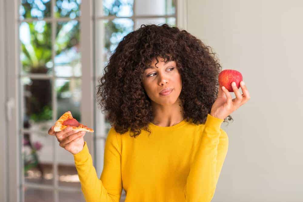 A woman wondering if she will choose the pizza or the apple.