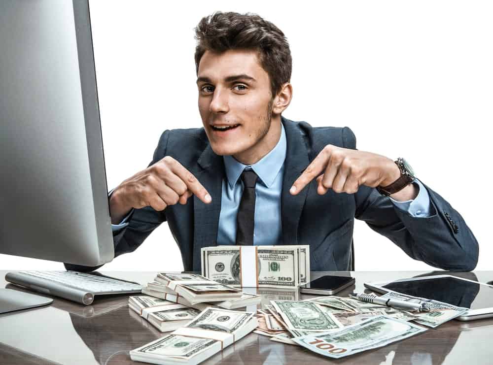 Businessman happily points at the wads of cash in front of a computer.