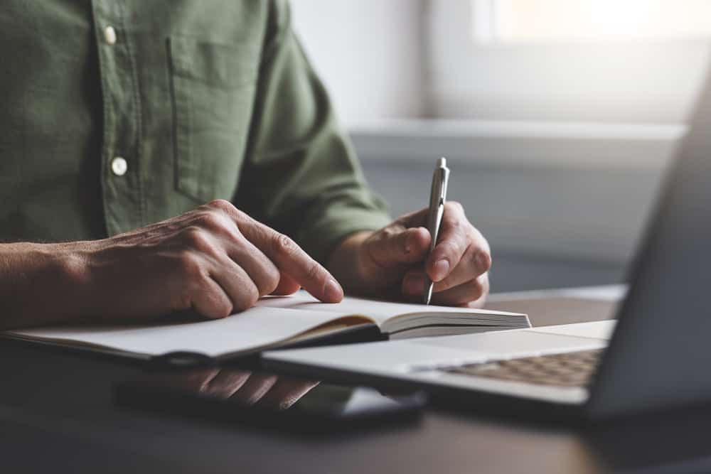 Cropped photo of a man writing on a notepad in front of an open laptop.